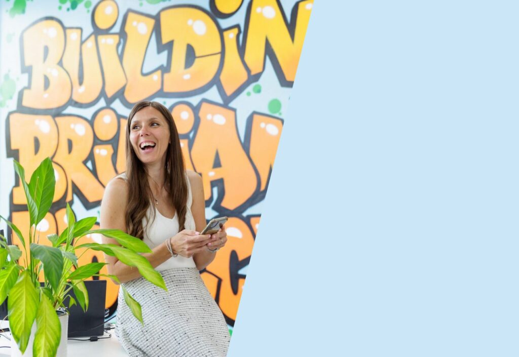 Building brilliant culture for Biotchs: Image shows a Singular employee with long brown hair laughing at something off camera. Behind her is a wall with the words "Building brilliant culture" graffitied onto it.
