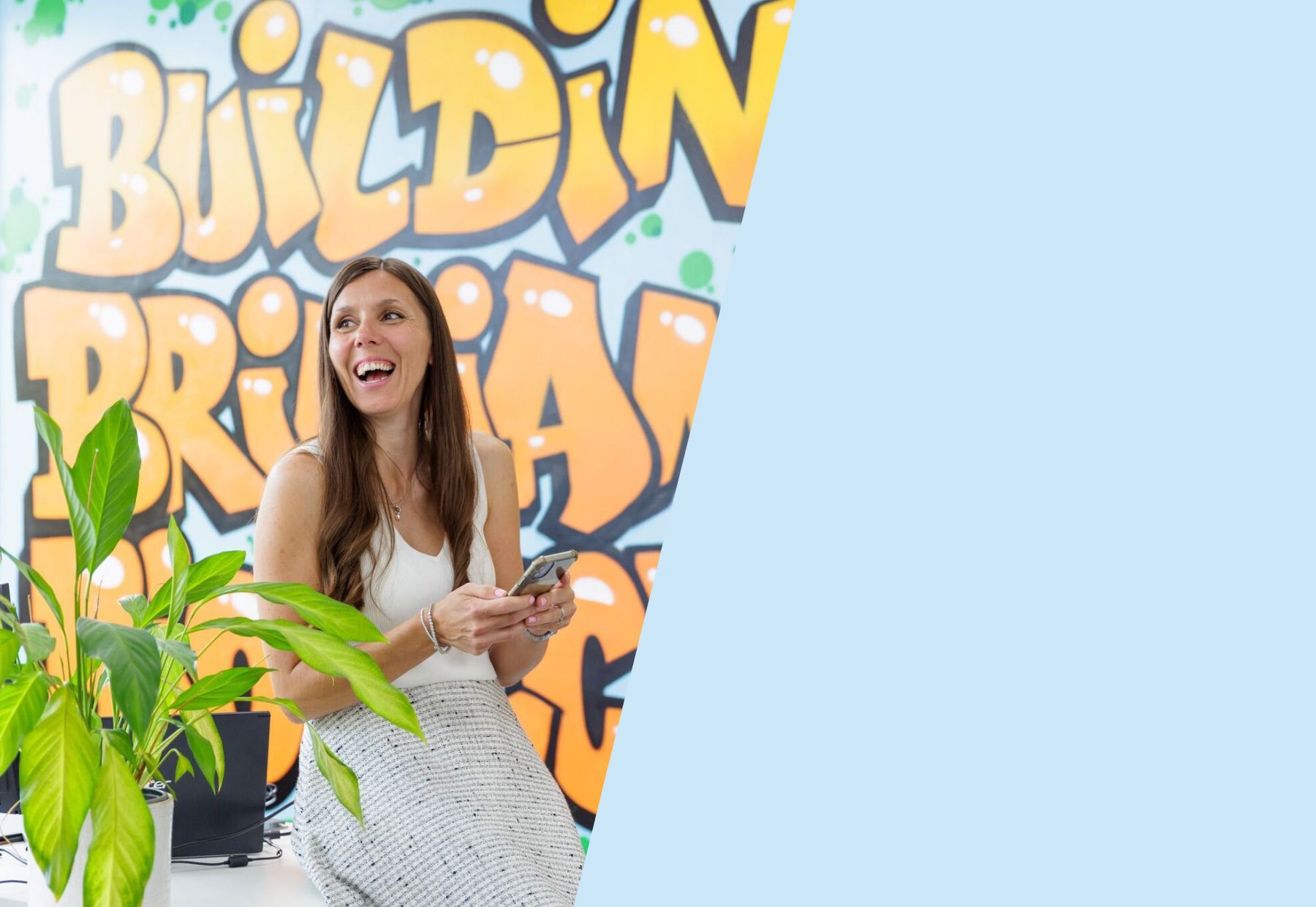 Building brilliant culture for Biotchs: Image shows a Singular employee with long brown hair laughing at something off camera. Behind her is a wall with the words "Building brilliant culture" graffitied onto it.